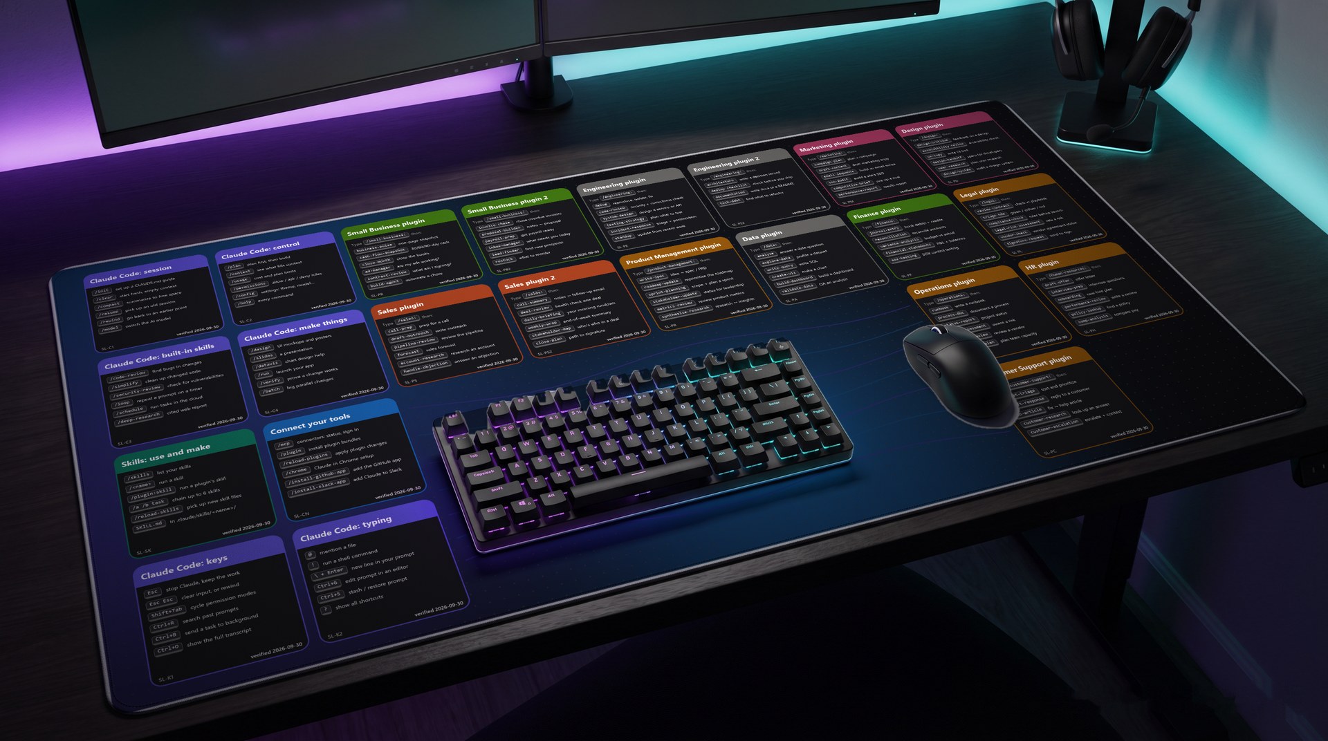 Mockup of the desk mat on a dark gaming desk lit purple and teal: an extended mat covered in colorful AI shortcut stickers, with a black mechanical keyboard in the middle, a black mouse on the right, two monitors and a headset stand behind.